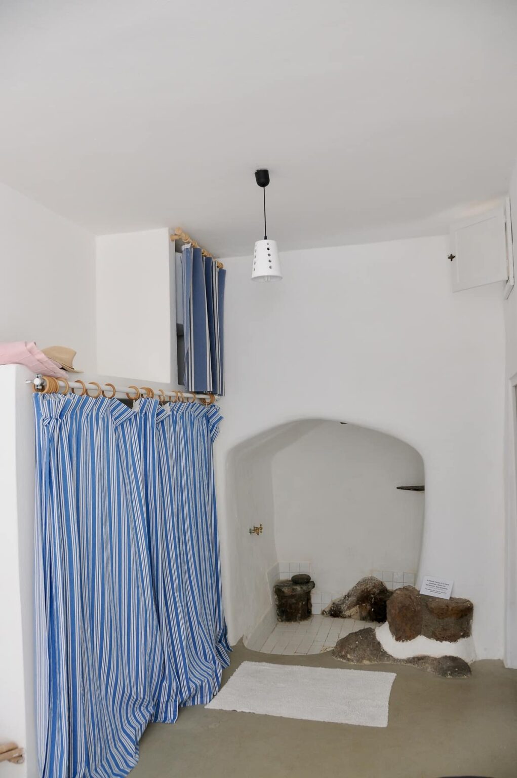 Interior view of a minimalist room featuring blue and white striped curtains, a hanging light fixture, and a small alcove with a stone wall and a faucet, designed for a cozy and functional space. | Only on SopranoVillas