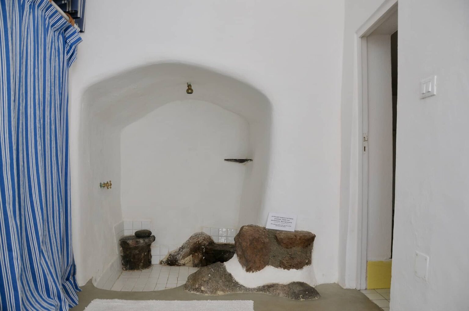 Interior view of a minimalist room featuring a white wall with an arched niche, decorative stones, and a blue and white striped curtain, highlighting simple design elements. | Only on SopranoVillas