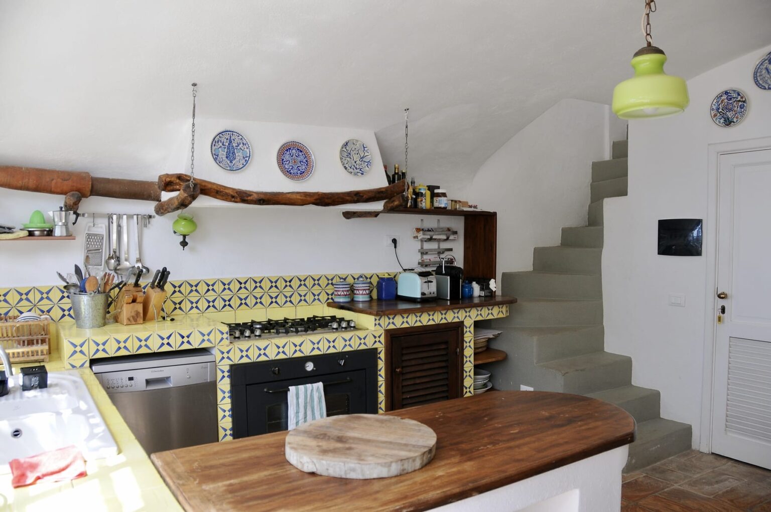 Modern kitchen interior featuring a wooden countertop, colorful tiled backsplash, and decorative plates on the wall. A staircase leads to the upper level, and a bright yellow pendant light adds a pop of color. | Only on SopranoVillas