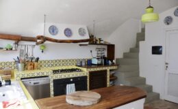 Modern kitchen interior featuring a wooden countertop, colorful tiled backsplash, and decorative plates on the wall. A staircase leads to the upper level, and a bright yellow pendant light adds a pop of color.