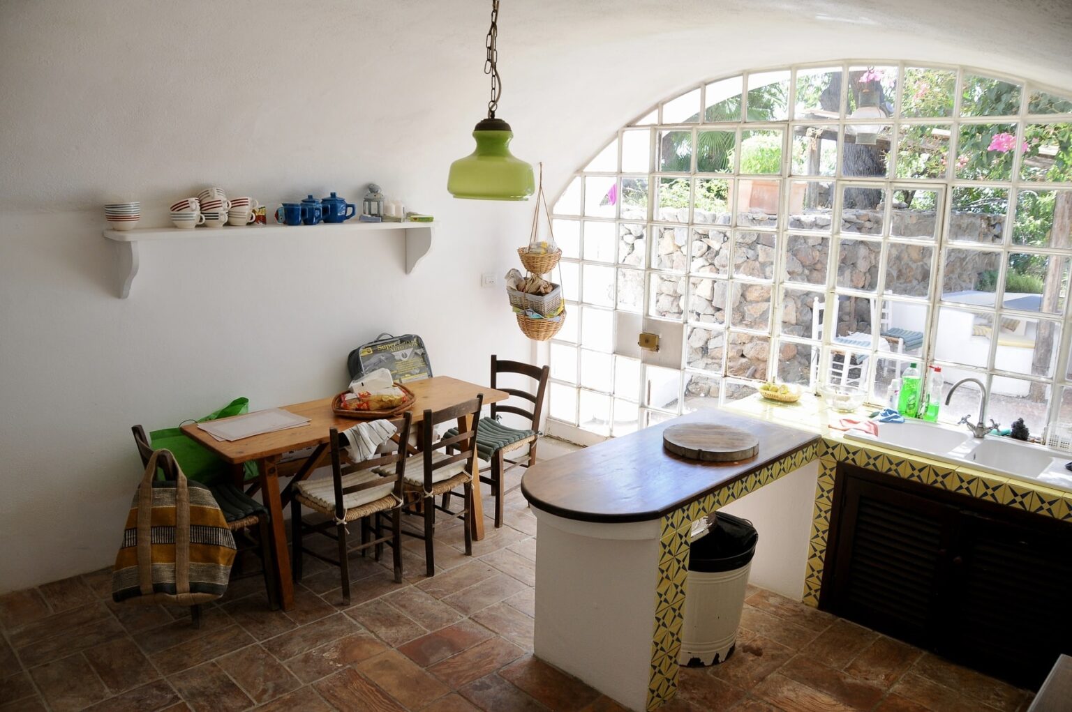 Cozy kitchen interior featuring a wooden dining table with chairs, a bright green pendant light, and a window with stone wall view. The shelf displays colorful dishware, while the kitchen area includes a sink and tiled countertop. Natural light enhances the inviting atmosphere. | Only on SopranoVillas