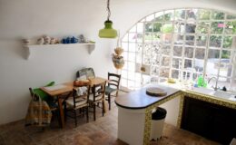 Cozy kitchen interior featuring a wooden dining table with chairs, a bright green pendant light, and a window with stone wall view. The shelf displays colorful dishware, while the kitchen area includes a sink and tiled countertop. Natural light enhances the inviting atmosphere.