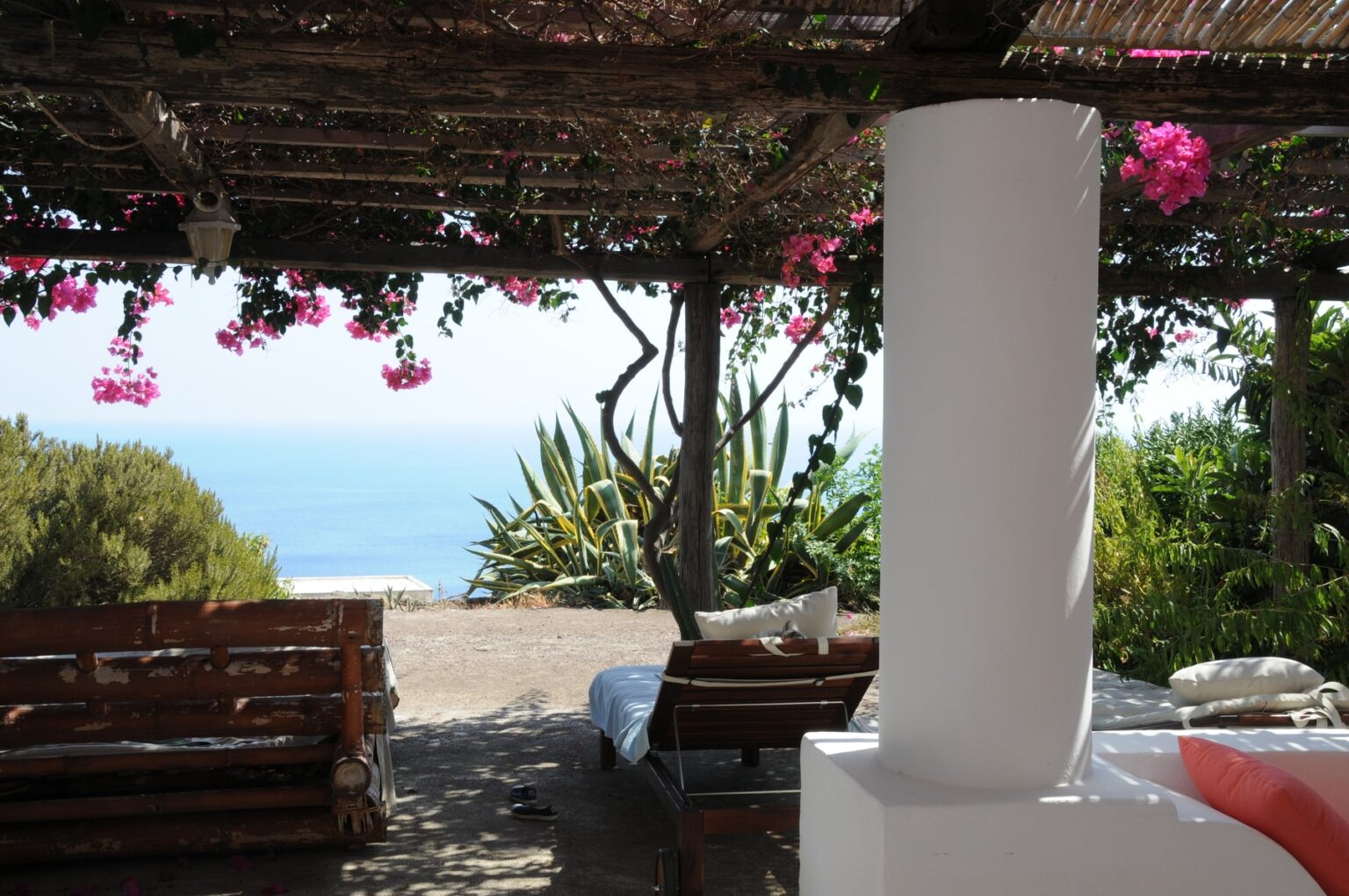 Cozy outdoor seating area under a pergola adorned with vibrant pink flowers, overlooking a serene ocean view. Ideal for relaxation and enjoying nature. | Only on SopranoVillas