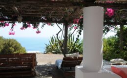 Cozy outdoor seating area under a pergola adorned with vibrant pink flowers, overlooking a serene ocean view. Ideal for relaxation and enjoying nature.
