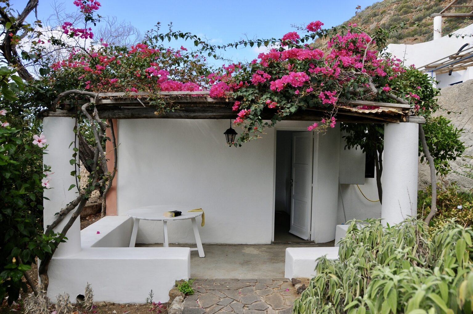Charming white cottage with a wooden roof adorned with vibrant pink bougainvillea flowers, surrounded by lush greenery and a clear blue sky. Perfect for a serene getaway or rustic vacation retreat. | Only on SopranoVillas