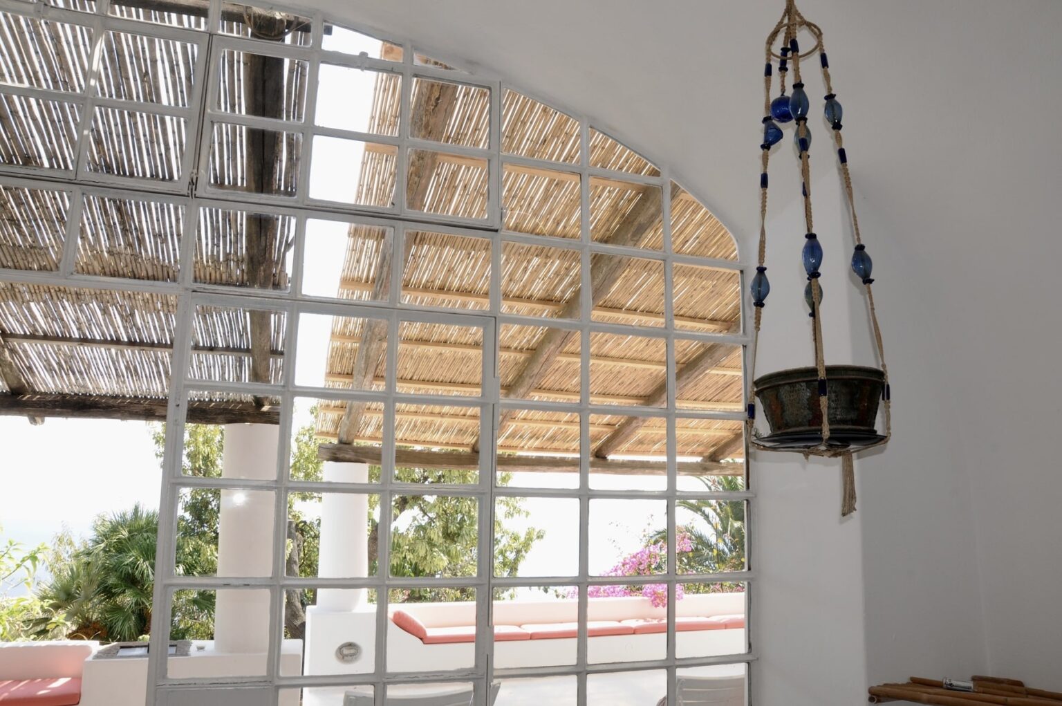 Interior view showcasing a whitewashed wall with a grid window, allowing natural light to filter in. A rustic thatched roof is visible outside, complemented by a hanging decorative planter. The scene features lush greenery and a hint of pink flowering plants, creating a serene and inviting atmosphere. Ideal for showcasing modern architecture and natural elements. | Only on SopranoVillas