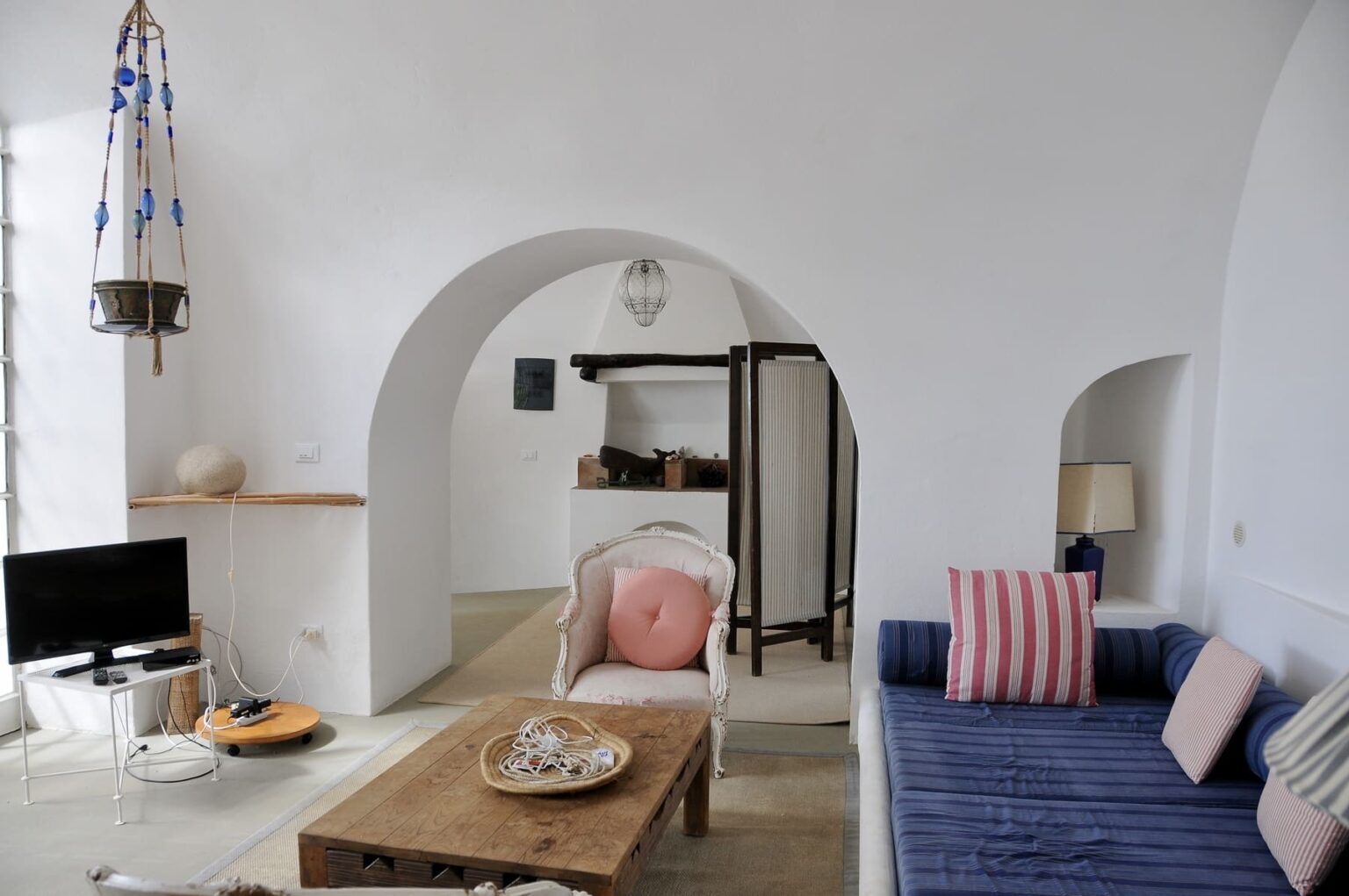 Cozy living room interior featuring white walls, an arched doorway, a wooden coffee table with decorative items, a blue sofa with pink striped cushions, and a small television. Natural light enhances the inviting atmosphere. | Only on SopranoVillas