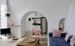 Cozy living room interior featuring white walls, an arched doorway, a wooden coffee table with decorative items, a blue sofa with pink striped cushions, and a small television. Natural light enhances the inviting atmosphere.
