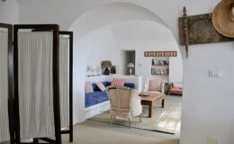 Cozy interior of a modern living room featuring a blue sofa, wicker chairs, decorative wall art, and a light-colored rug, set in a bright, airy space with arched ceilings and a room divider.