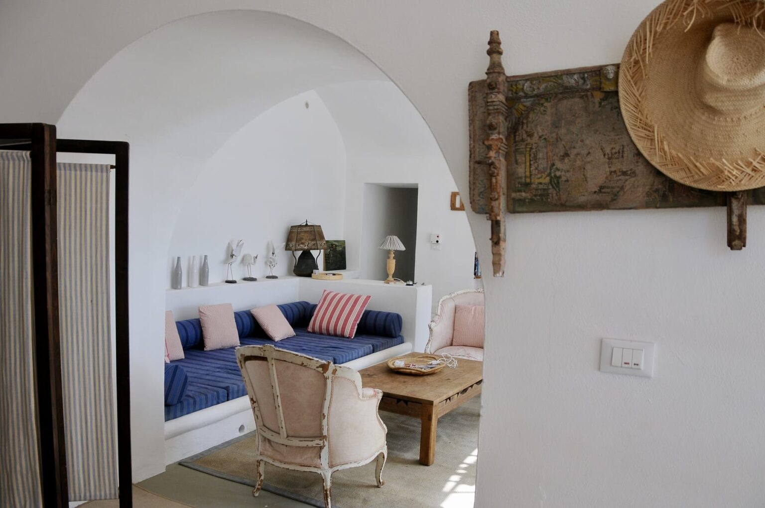 Bright and airy living room featuring a cozy blue sofa with striped pillows, a wooden coffee table, and stylish decor. The room has white walls and an arched doorway, creating a welcoming atmosphere. A straw hat and decorative wall piece add a touch of charm. | Only on SopranoVillas