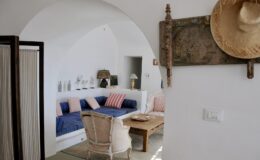 Bright and airy living room featuring a cozy blue sofa with striped pillows, a wooden coffee table, and stylish decor. The room has white walls and an arched doorway, creating a welcoming atmosphere. A straw hat and decorative wall piece add a touch of charm.