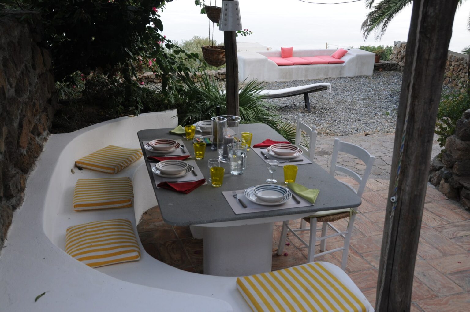 Outdoor dining area with a stone wall, featuring a gray table set with colorful plates, glasses, and napkins. Yellow striped cushions provide seating, surrounded by lush greenery and a view of a distant lounge area with a pink sofa. Ideal for al fresco dining and relaxation. | Only on SopranoVillas