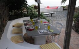Outdoor dining area with a stone wall, featuring a gray table set with colorful plates, glasses, and napkins. Yellow striped cushions provide seating, surrounded by lush greenery and a view of a distant lounge area with a pink sofa. Ideal for al fresco dining and relaxation.