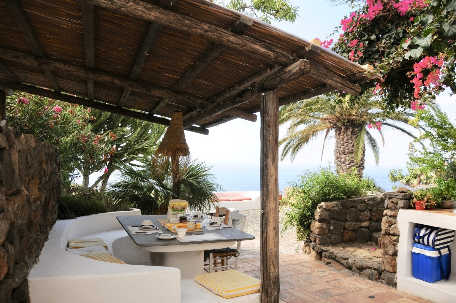Outdoor dining area with a stone wall, white seating, and a table set for breakfast, surrounded by lush greenery and blooming flowers, overlooking a scenic ocean view. | Only on SopranoVillas