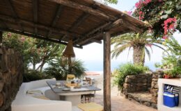 Outdoor dining area with a stone wall, white seating, and a table set for breakfast, surrounded by lush greenery and blooming flowers, overlooking a scenic ocean view.