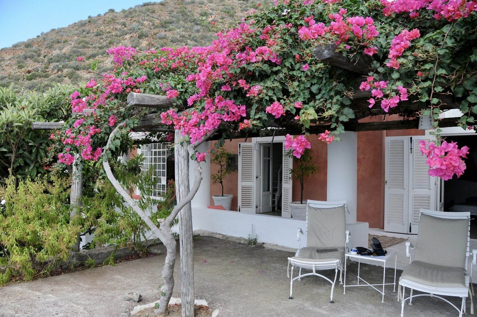 Charming outdoor patio with white chairs under a vibrant bougainvillea canopy, set against a backdrop of a rustic house and lush greenery in a mountainous landscape. | Only on SopranoVillas