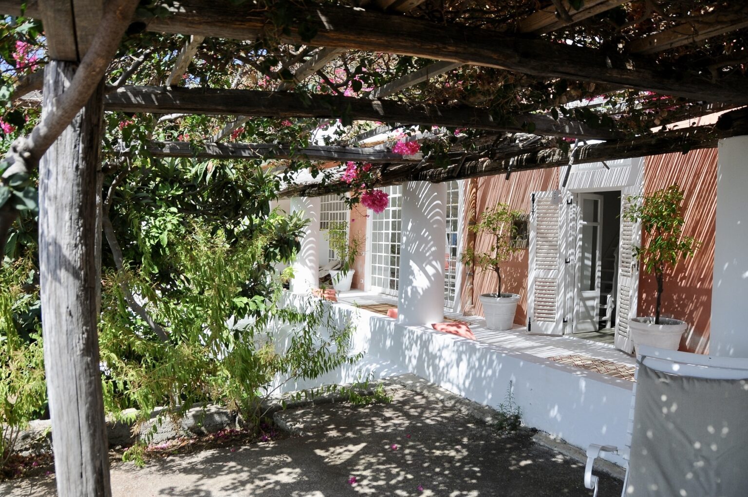 Charming outdoor patio with a wooden pergola adorned with blooming flowers, surrounded by greenery and a white-walled house, creating a serene and inviting atmosphere. | Only on SopranoVillas