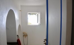 Interior view of a bright, minimalist bathroom featuring a window, a gold shower fixture, and a red towel hanging nearby, emphasizing a clean and airy atmosphere.