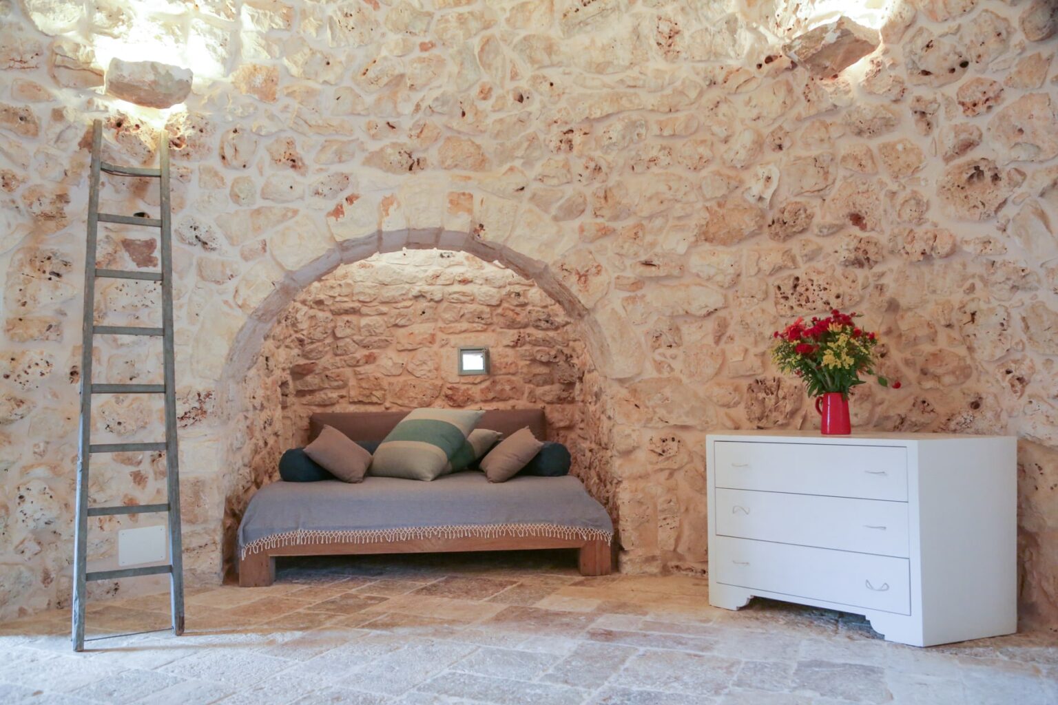 Cozy interior of a rustic stone room featuring a comfortable daybed with decorative pillows, a white dresser, and a wooden ladder, illuminated by natural light. | Only on SopranoVillas