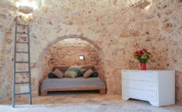 Cozy interior of a rustic stone room featuring a comfortable daybed with decorative pillows, a white dresser, and a wooden ladder, illuminated by natural light.