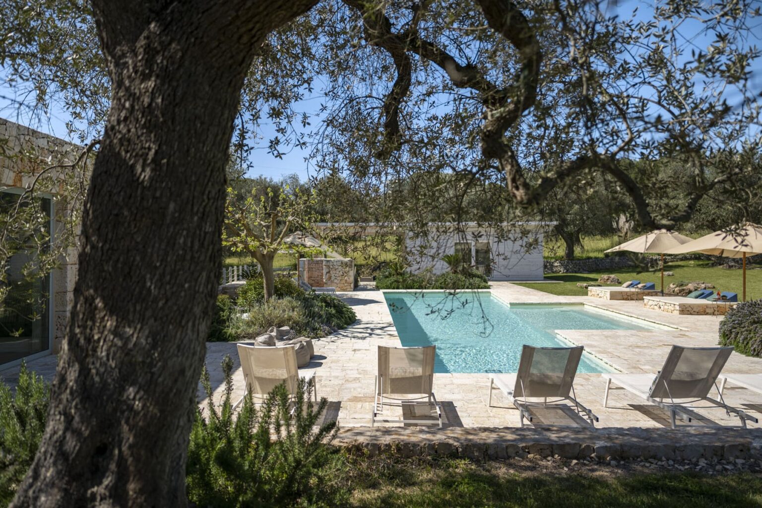 Luxurious outdoor pool area surrounded by greenery, featuring lounge chairs, sun umbrellas, and a serene landscape in a sunny setting. | Only on SopranoVillas