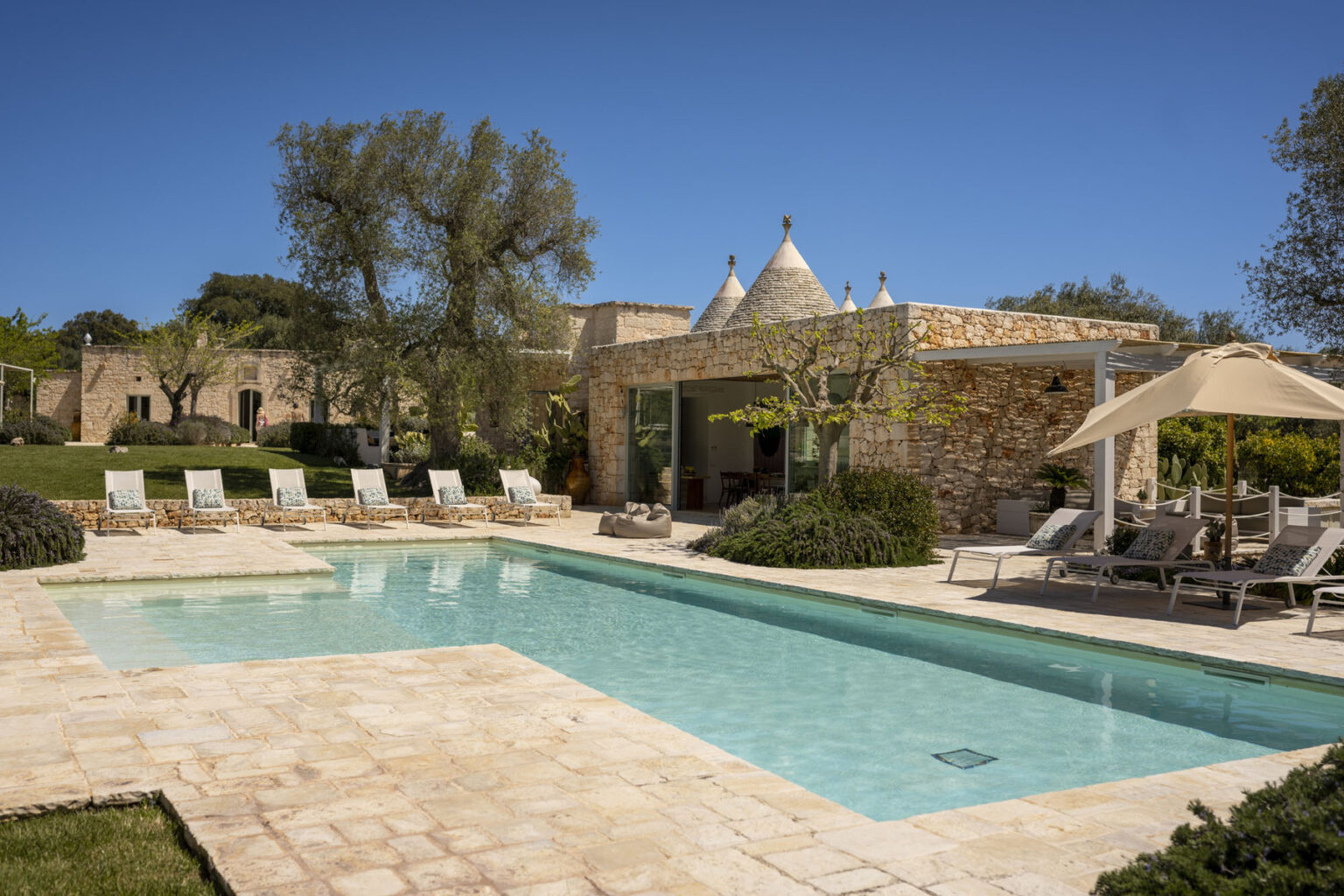 Luxury villa with a swimming pool, surrounded by sun loungers and lush greenery, featuring a stone facade and a clear blue sky. Ideal for relaxation and outdoor living. | Only on SopranoVillas