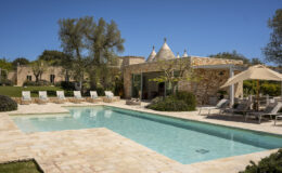 Luxury villa with a swimming pool, surrounded by sun loungers and lush greenery, featuring a stone facade and a clear blue sky. Ideal for relaxation and outdoor living.