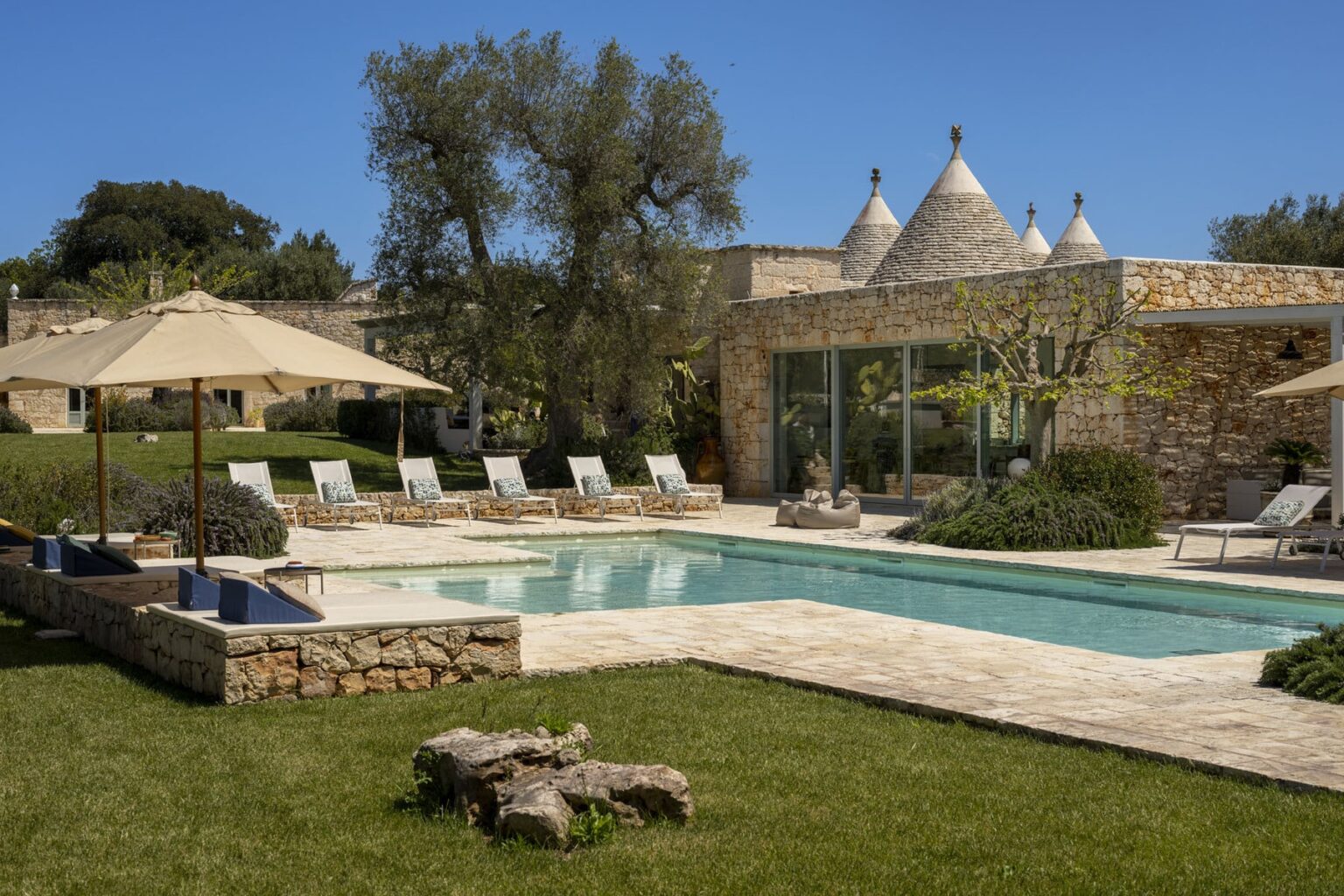 Luxury villa with a swimming pool, surrounded by sun loungers and shaded by umbrellas, featuring traditional trulli architecture in a lush green landscape. Ideal for relaxation and outdoor living. | Only on SopranoVillas