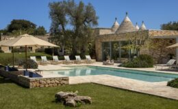 Luxury villa with a swimming pool, surrounded by sun loungers and shaded by umbrellas, featuring traditional trulli architecture in a lush green landscape. Ideal for relaxation and outdoor living.