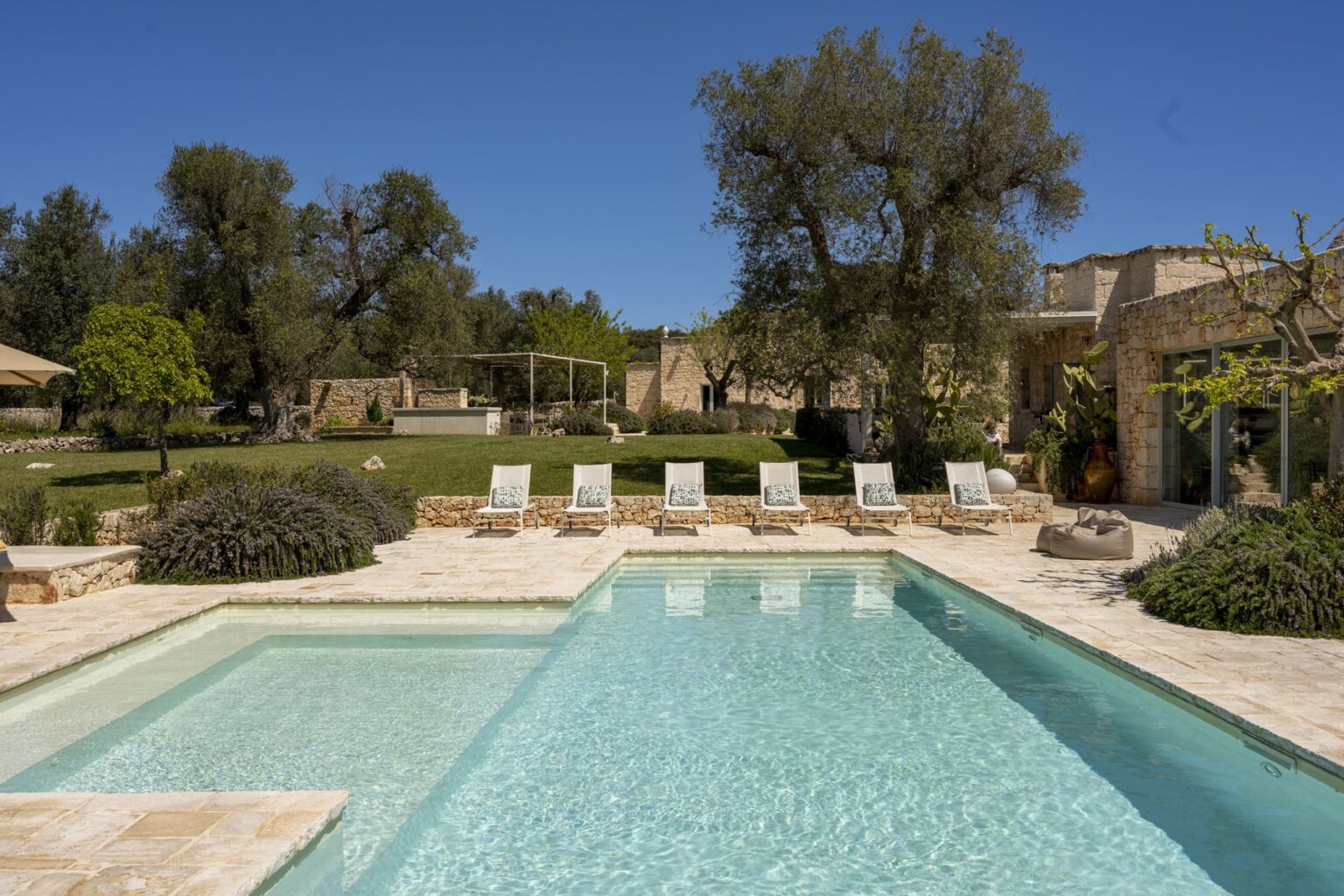 Luxurious outdoor swimming pool surrounded by lounge chairs, set in a beautifully landscaped garden with trees and stone architecture under a clear blue sky. Ideal for relaxation and summer retreats. | Only on SopranoVillas