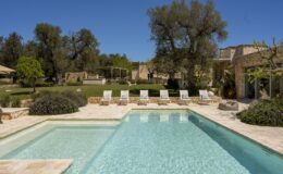 Luxurious outdoor swimming pool surrounded by lounge chairs, set in a beautifully landscaped garden with trees and stone architecture under a clear blue sky. Ideal for relaxation and summer retreats.