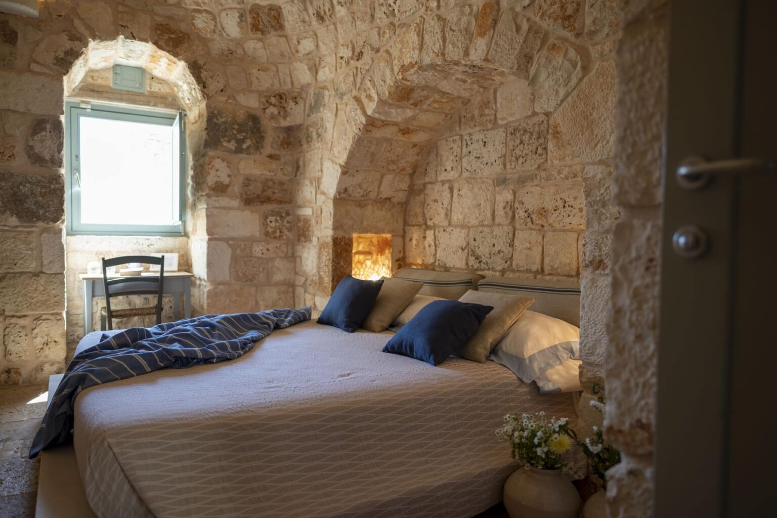Cozy bedroom in a stone-walled setting featuring a large bed with blue pillows, a wooden chair, and a window providing natural light, ideal for a relaxing stay. | Only on SopranoVillas
