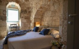 Cozy bedroom in a stone-walled setting featuring a large bed with blue pillows, a wooden chair, and a window providing natural light, ideal for a relaxing stay.