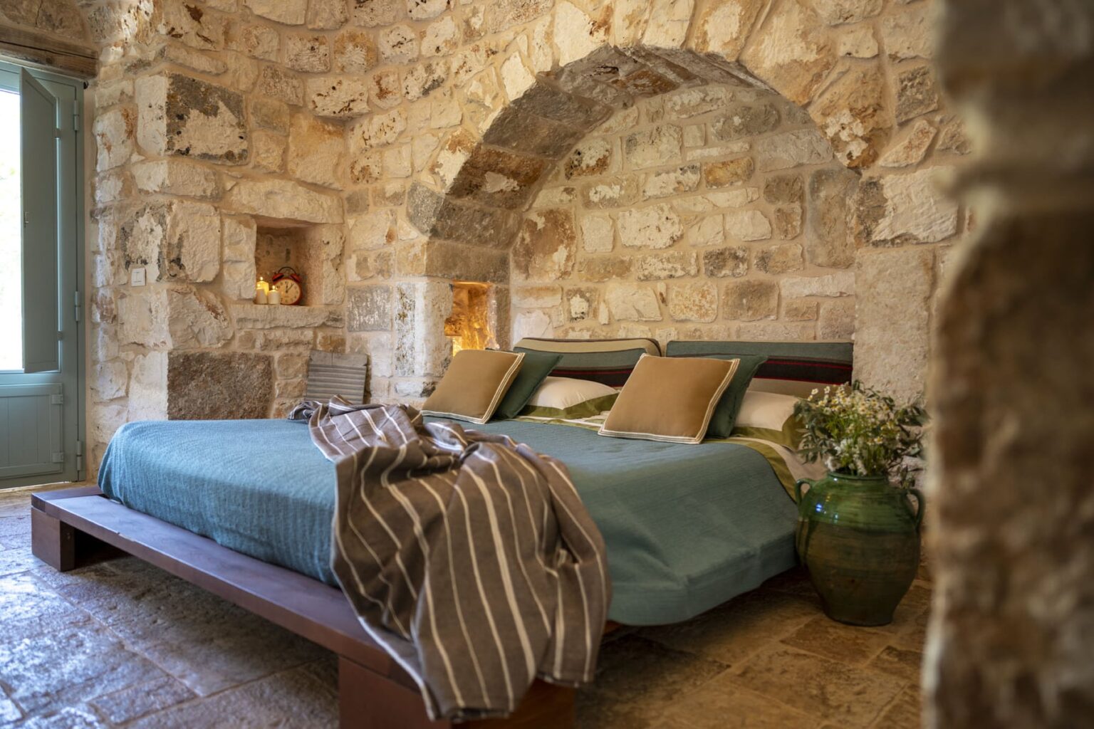Cozy bedroom featuring a stone wall design, two beds with teal bedding, decorative pillows, and a rustic wooden bed frame. Natural light streams through a window, enhancing the warm, inviting atmosphere. A green vase with flowers adds a touch of nature to the decor. | Only on SopranoVillas