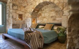 Cozy bedroom featuring a stone wall design, two beds with teal bedding, decorative pillows, and a rustic wooden bed frame. Natural light streams through a window, enhancing the warm, inviting atmosphere. A green vase with flowers adds a touch of nature to the decor.