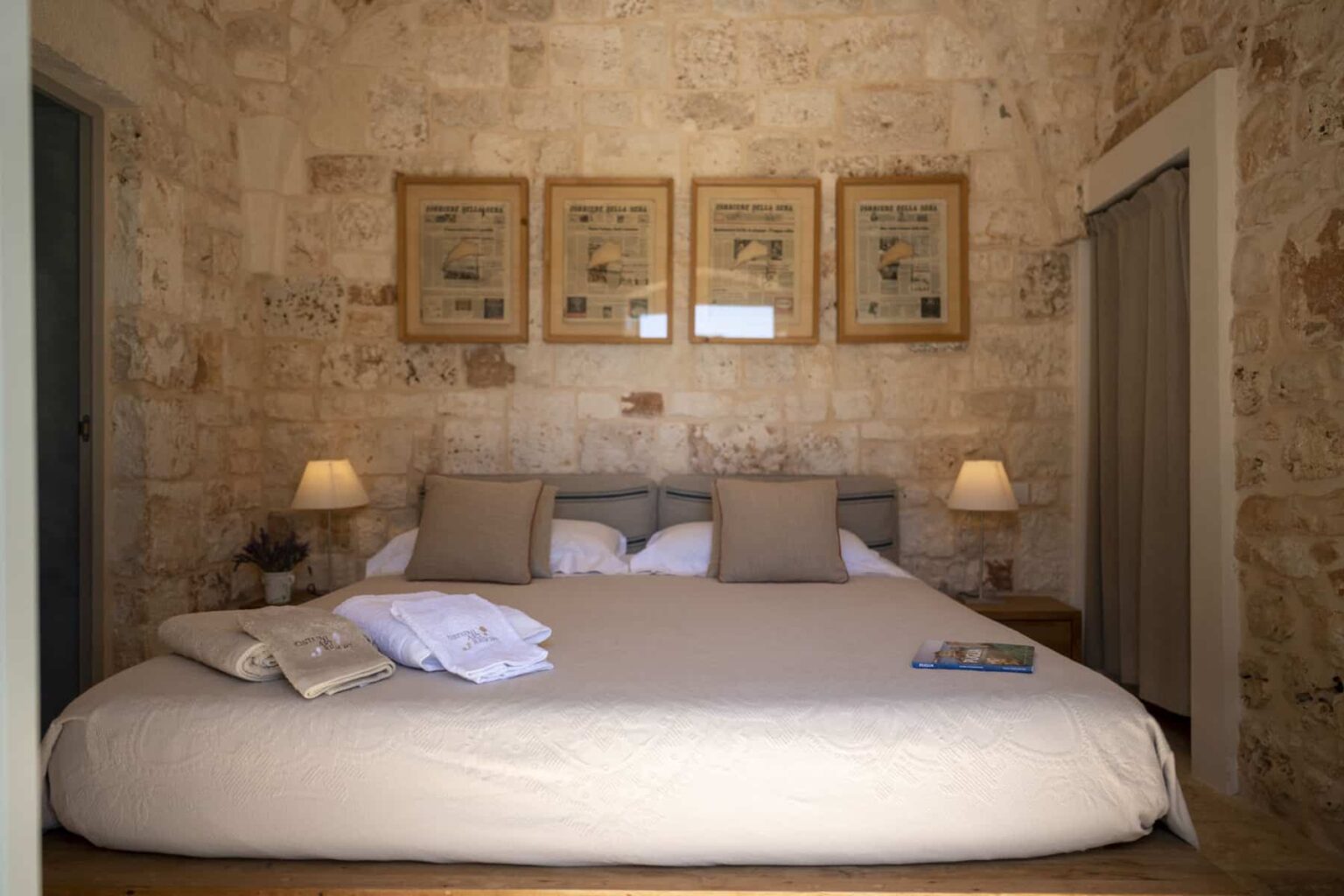 Cozy bedroom featuring a large bed with beige linens, two bedside lamps, and stone wall decor. Framed artwork hangs above the bed, adding a touch of elegance to the serene atmosphere. | Only on SopranoVillas