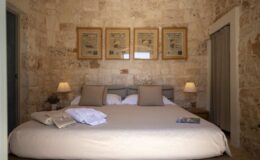 Cozy bedroom featuring a large bed with beige linens, two bedside lamps, and stone wall decor. Framed artwork hangs above the bed, adding a touch of elegance to the serene atmosphere.