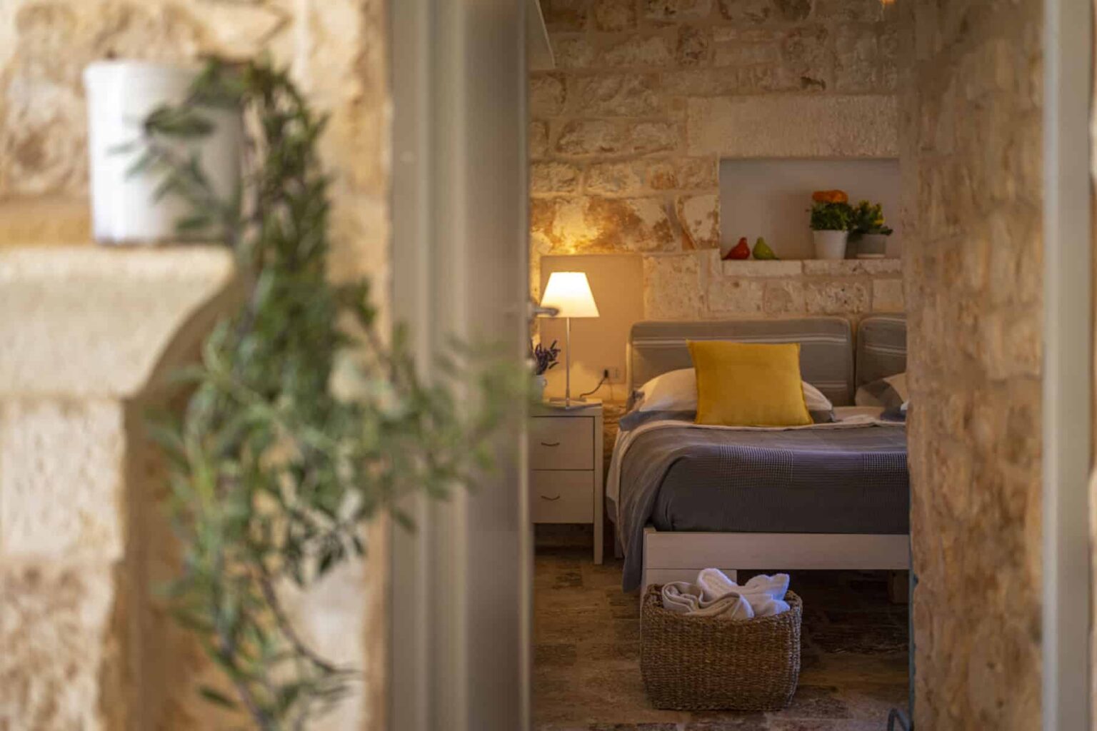 Cozy bedroom with stone walls, featuring a comfortable bed with gray bedding and a bright yellow pillow, soft lighting, and decorative plants, creating a warm and inviting atmosphere. | Only on SopranoVillas