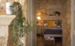 Cozy bedroom with stone walls, featuring a comfortable bed with gray bedding and a bright yellow pillow, soft lighting, and decorative plants, creating a warm and inviting atmosphere.