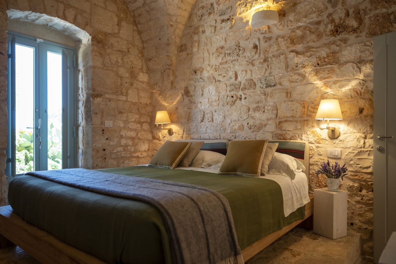 Cozy bedroom featuring a stone wall, a comfortable bed with green bedding and decorative pillows, and natural light streaming through blue-framed windows. The room is illuminated by wall sconces, and a small white bedside table holds a potted lavender plant, enhancing the inviting atmosphere. | Only on SopranoVillas