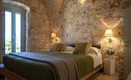 Cozy bedroom featuring a stone wall, a comfortable bed with green bedding and decorative pillows, and natural light streaming through blue-framed windows. The room is illuminated by wall sconces, and a small white bedside table holds a potted lavender plant, enhancing the inviting atmosphere.