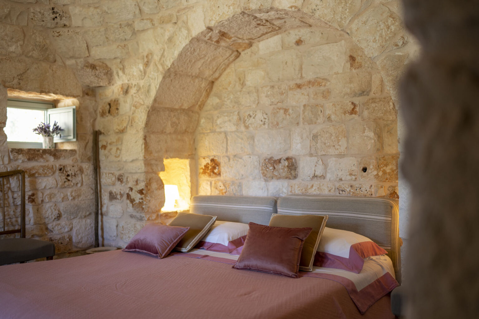 Cozy bedroom interior featuring a stone wall, arched architecture, a comfortable bed with decorative pillows, and a window with a vase of lavender, creating a warm and inviting atmosphere. | Only on SopranoVillas