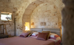Cozy bedroom interior featuring a stone wall, arched architecture, a comfortable bed with decorative pillows, and a window with a vase of lavender, creating a warm and inviting atmosphere.