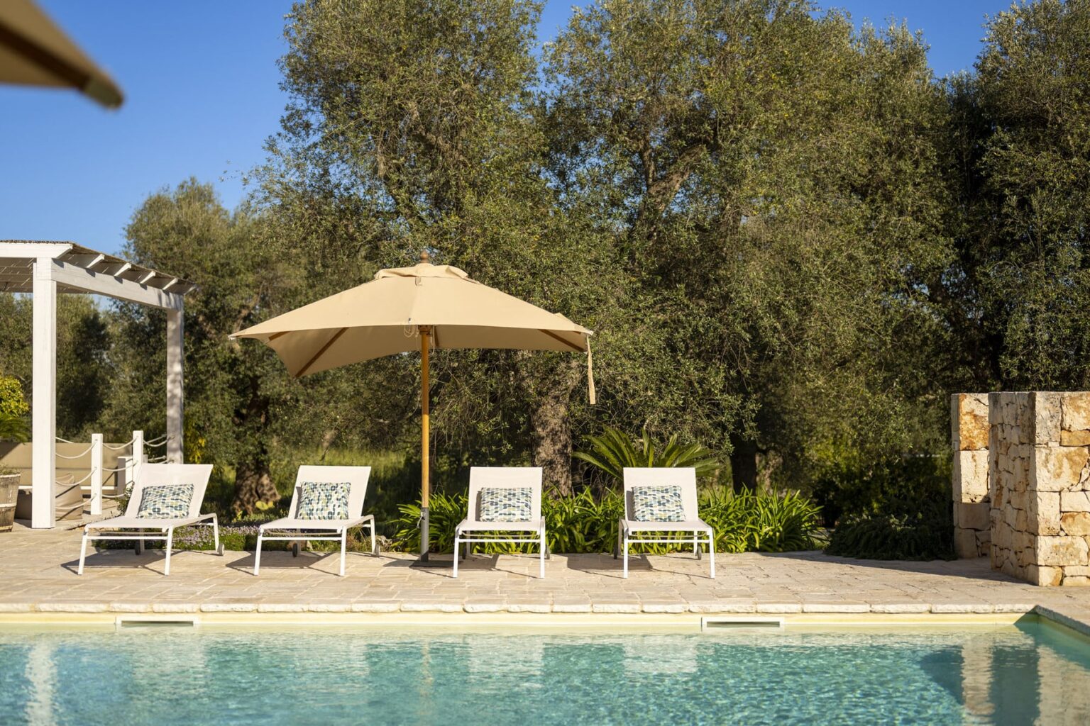 Inviting outdoor pool area featuring lounge chairs and a large umbrella, surrounded by lush greenery and olive trees, perfect for relaxation and leisure. | Only on SopranoVillas