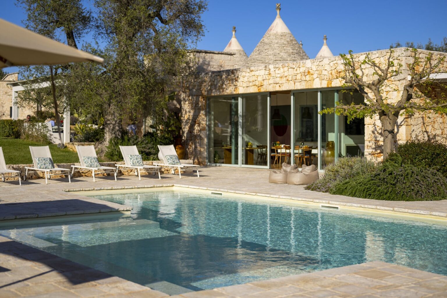 Modern villa with a swimming pool, surrounded by lush greenery and sun loungers, featuring stone architecture and large glass windows under a clear blue sky. | Only on SopranoVillas