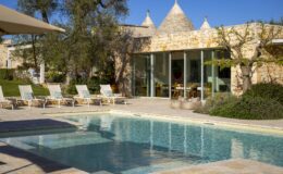 Modern villa with a swimming pool, surrounded by lush greenery and sun loungers, featuring stone architecture and large glass windows under a clear blue sky.
