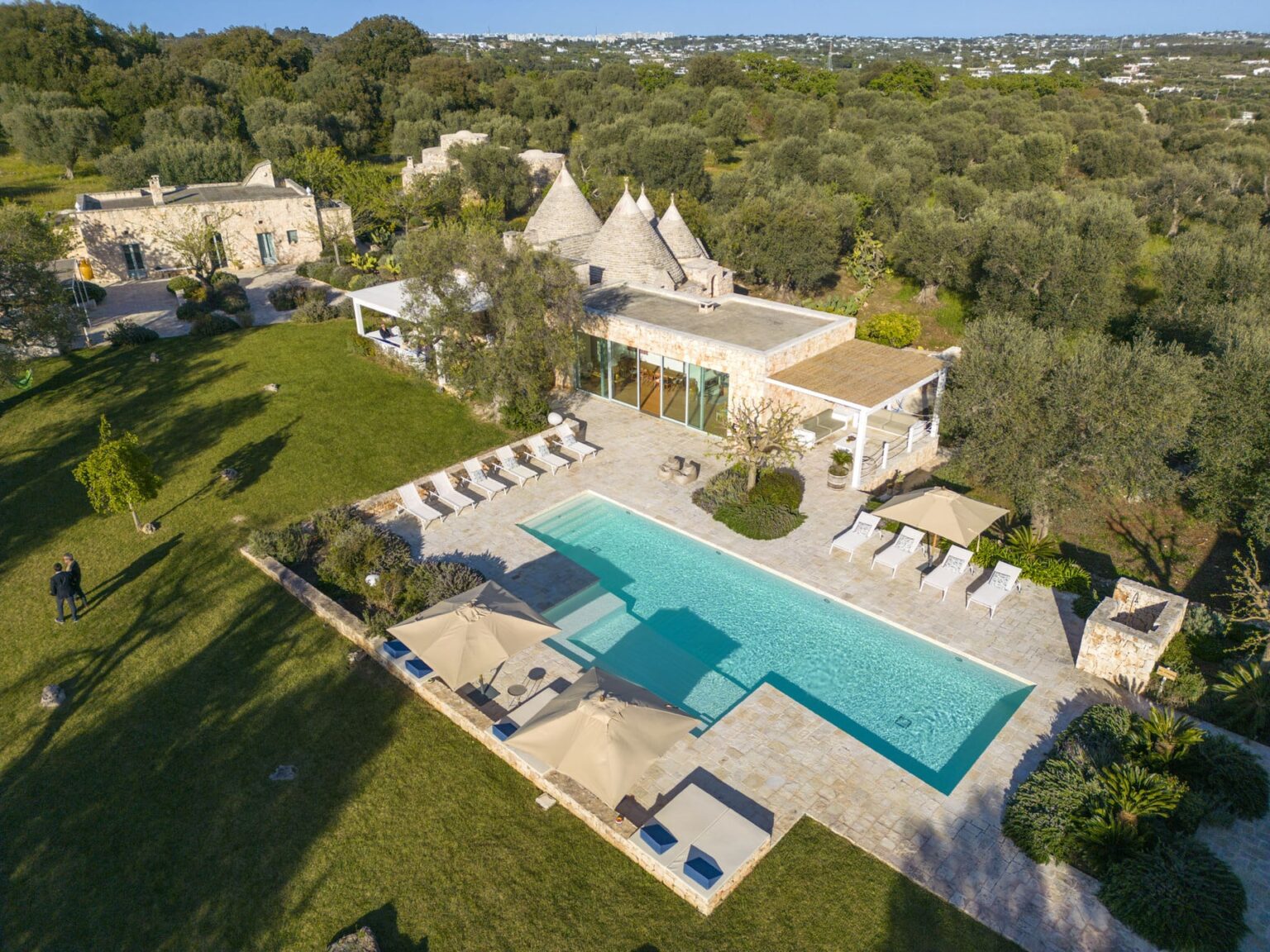 Aerial view of a luxurious villa featuring a large swimming pool surrounded by sun loungers, set in a lush green landscape with olive trees and traditional stone structures in the background. Ideal for vacation rentals or serene getaways. | Only on SopranoVillas