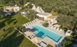 Aerial view of a luxurious villa featuring a large swimming pool surrounded by sun loungers, set in a lush green landscape with olive trees and traditional stone structures in the background. Ideal for vacation rentals or serene getaways.