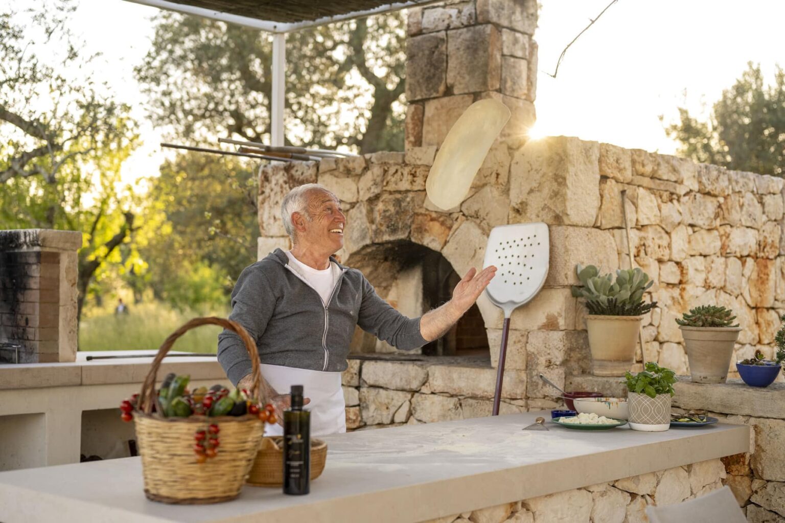 Smiling elderly man tossing pizza dough in an outdoor kitchen with stone walls, surrounded by cooking utensils, fresh ingredients, and a warm sunset ambiance. | Only on SopranoVillas