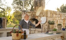 Smiling elderly man tossing pizza dough in an outdoor kitchen with stone walls, surrounded by cooking utensils, fresh ingredients, and a warm sunset ambiance.